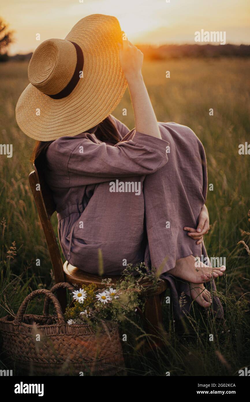 Beautiful woman in linen dress and straw hat sitting on rustic chair ...