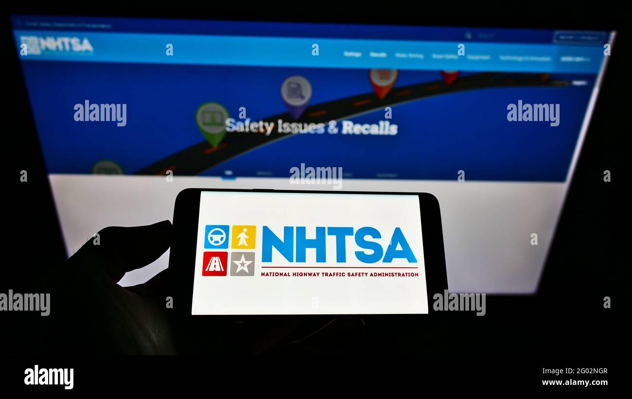 Person holding smartphone with logo of National Highway Traffic Safety Administration (NHTSA) on screen in front of website. Focus on phone display. Stock Photo
