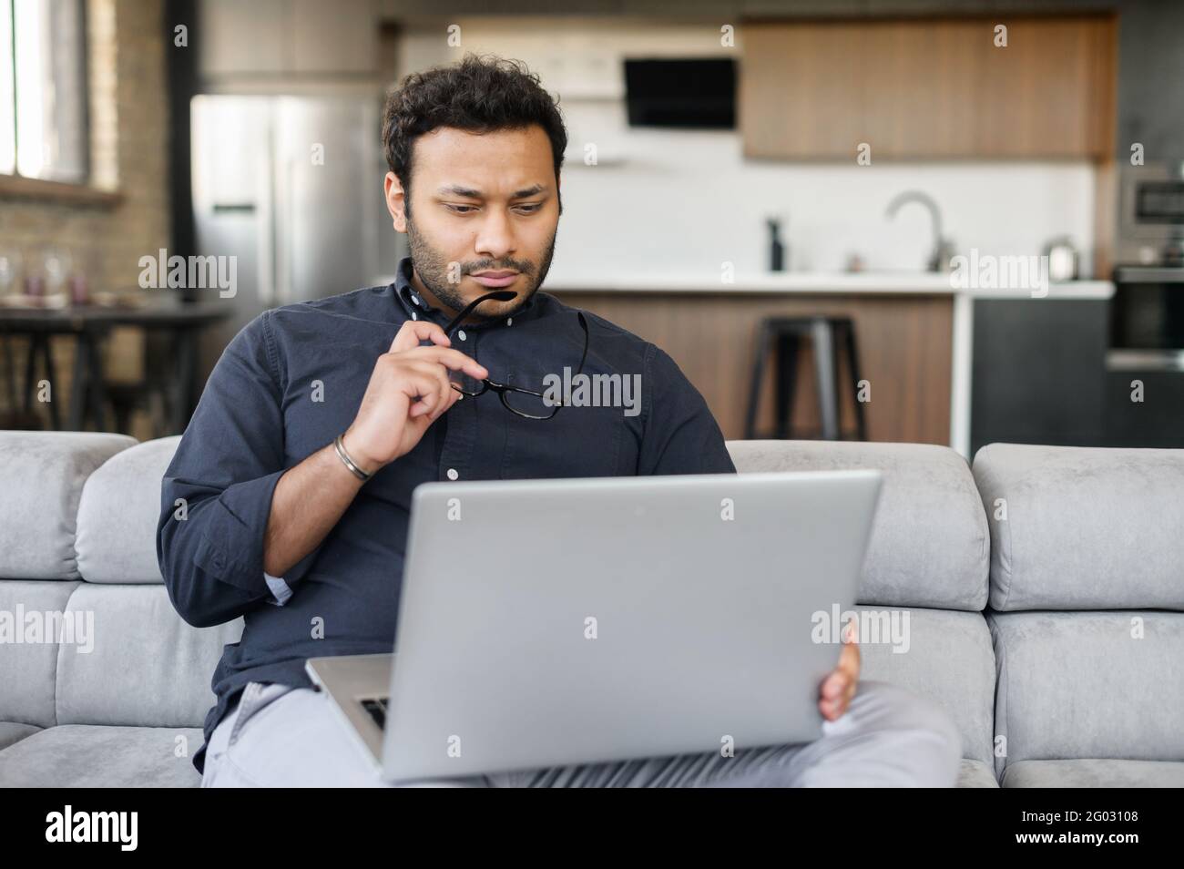Thoughtful indian man using laptop sitting on the couch at home, a hindu guy looks at the screen, looking for new sturtup idea, solving difficalt tasks, planning future Stock Photo