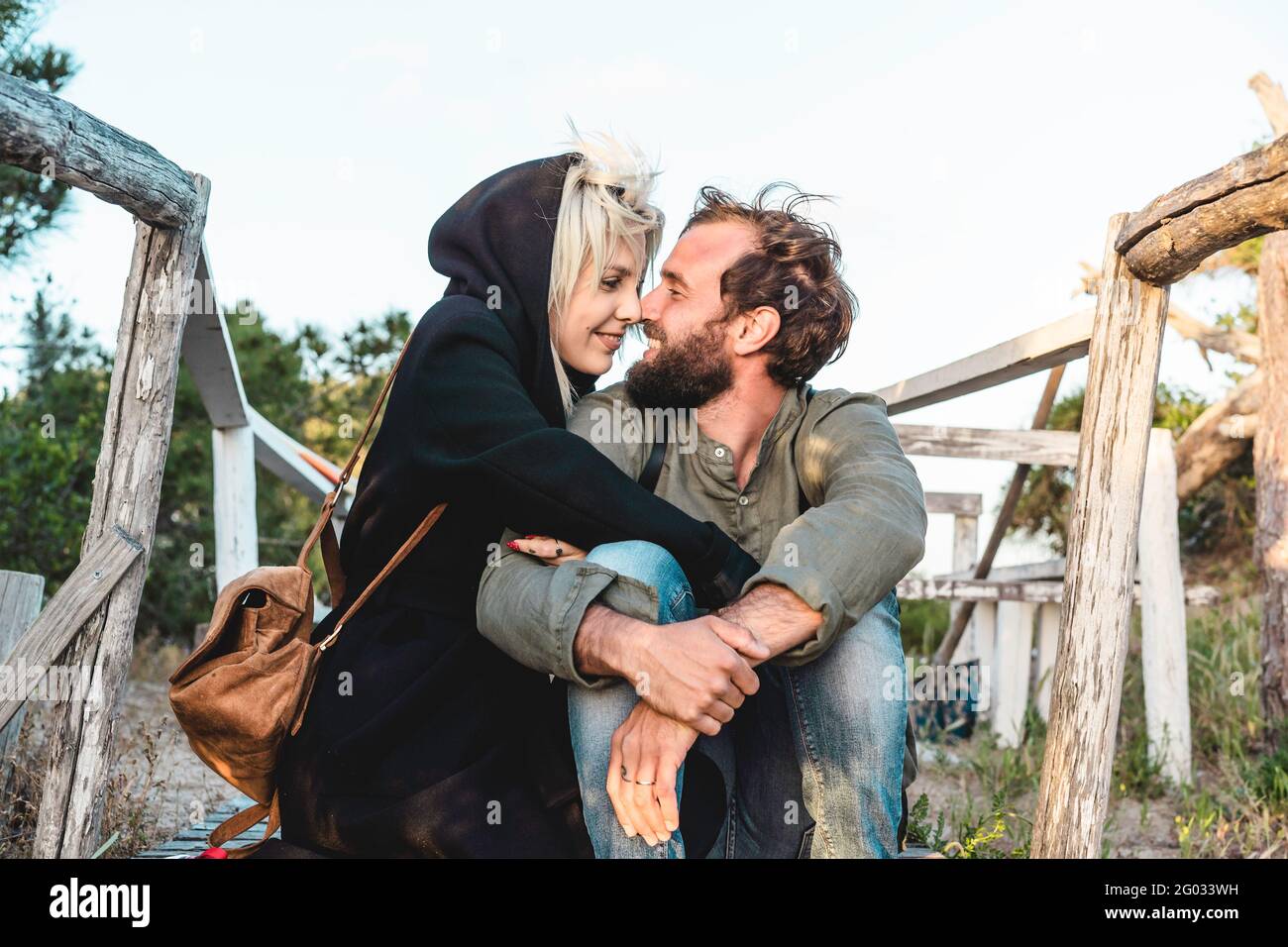 Romantic happy couple in love sitting on wooden stairs on dunes at the wild beach at sunset - Boyfriend and girlfriend looking into each other's eyes Stock Photo