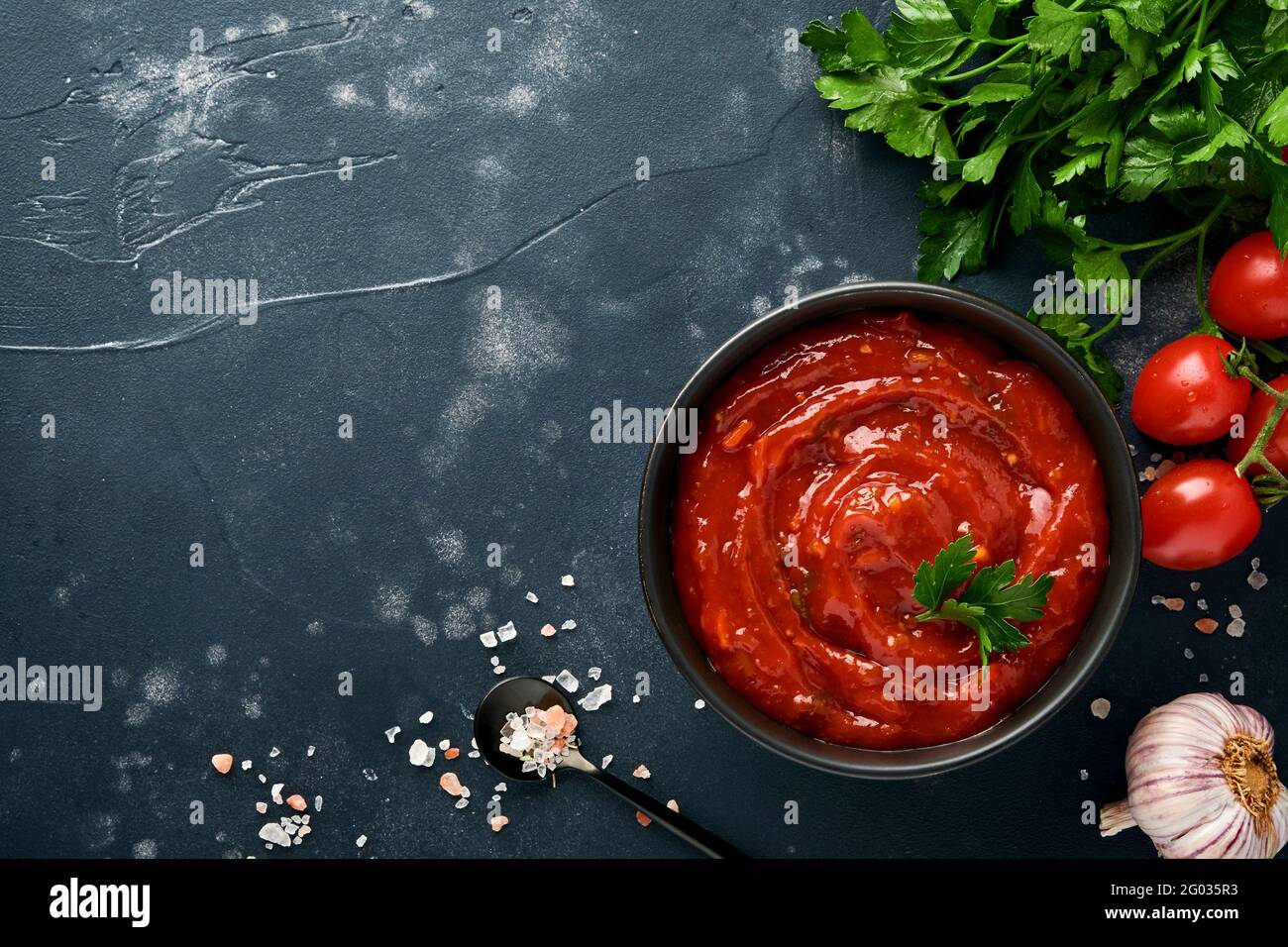 Tomato ketchup spicy sauce in a black bowl with spices and fresh tomatoes on a dark slate, stone or concrete background. Top view with copy space. Stock Photo