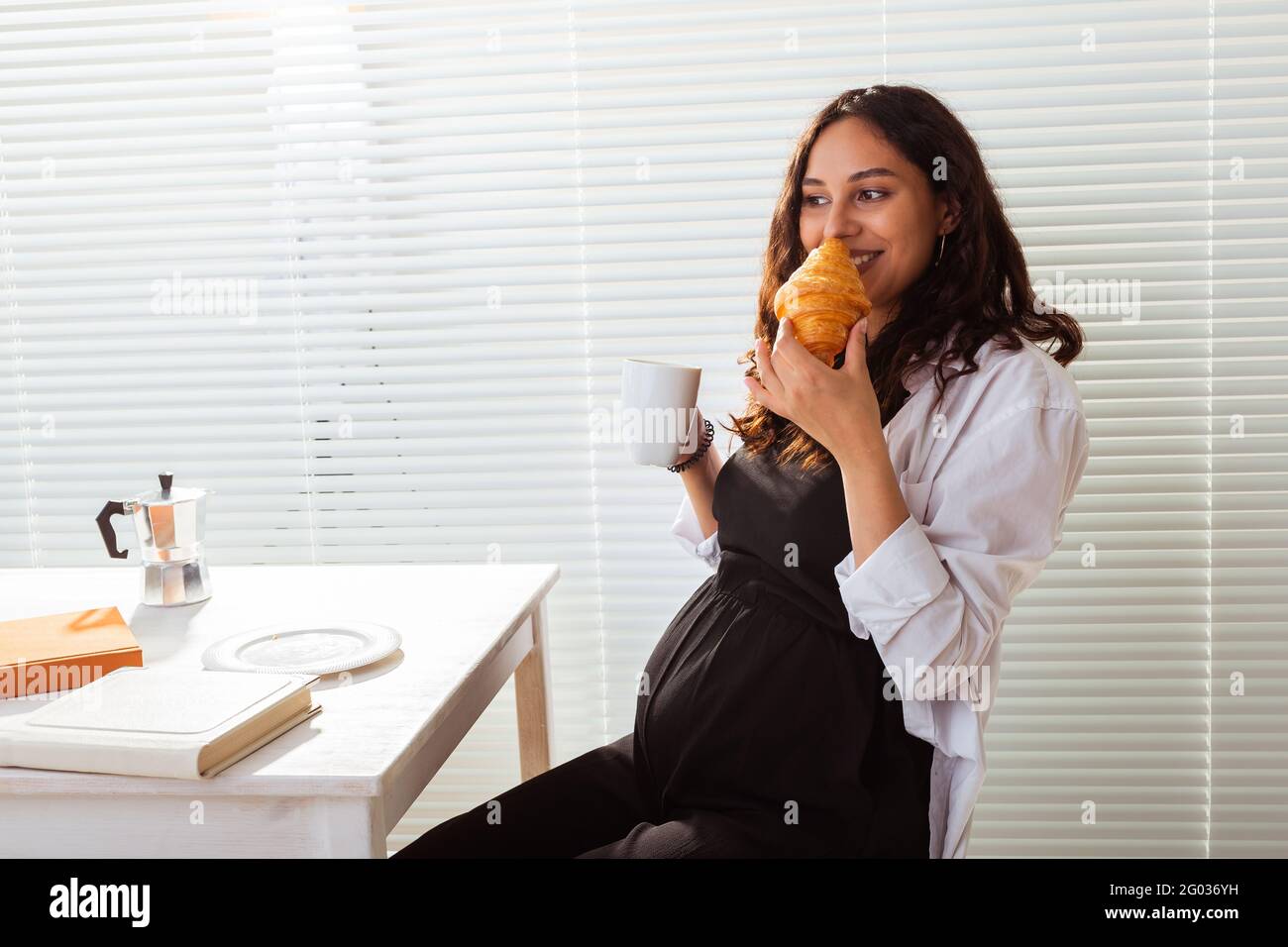 Happy pregnant young beautiful woman eating croissant during morning