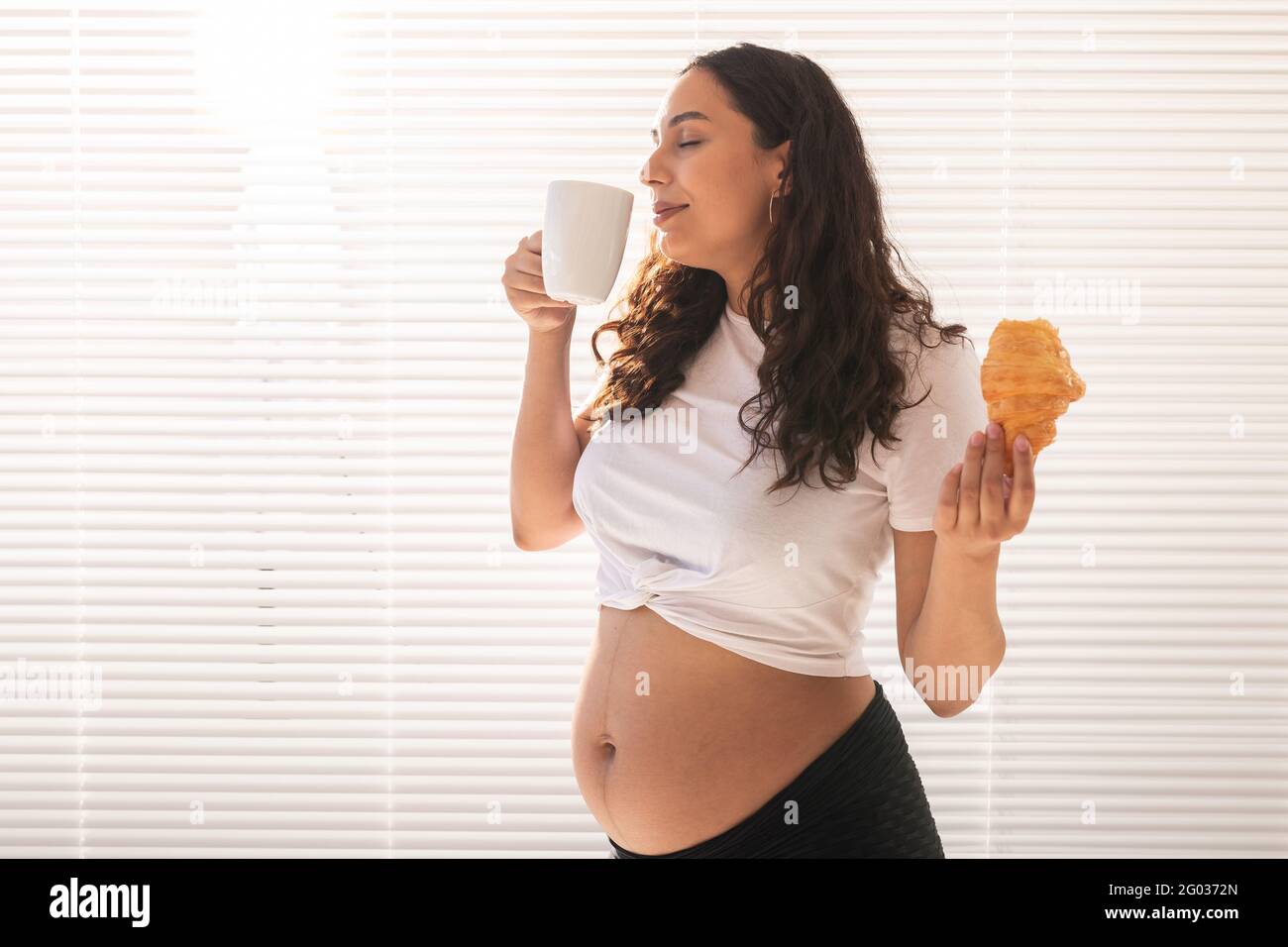 Beautiful pregnant woman holding croissant and cup of coffee in her ...