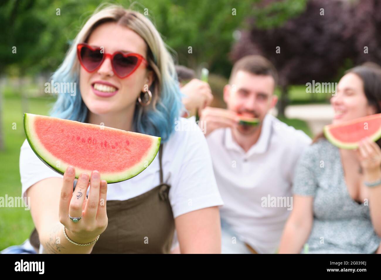 Happy young woman smiling holding a piece of watermelon and showing it to the camera, friends eating on the background. Stock Photo