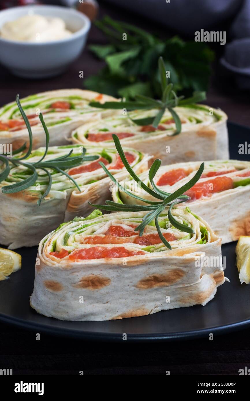 Rolls of thin pita bread and red salted salmon with lettuce leaves on a black ceramic plate, dark wooden background Stock Photo