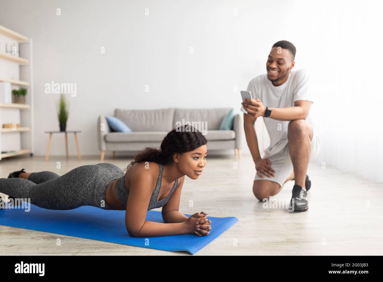 Millennial black couple training as team at home, athletic woman standing in plank pose, her boyfriend noting time Stock Photo