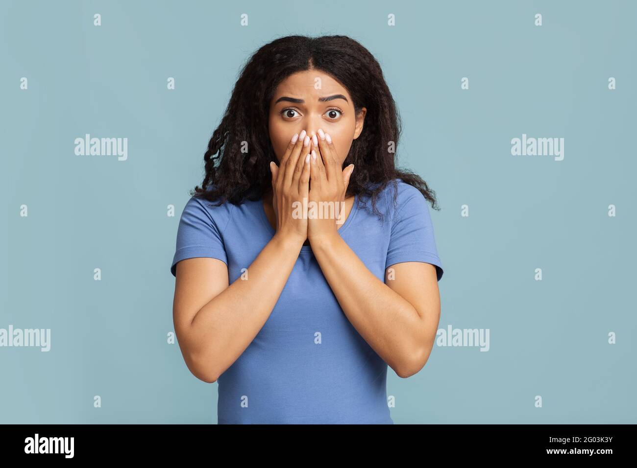 Oh no. Shocked and scared african american lady covering her mouth with palms, standing over blue studio background Stock Photo
