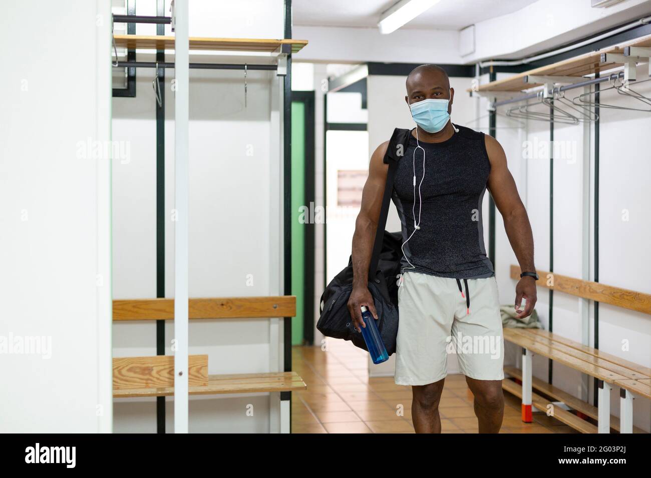 Young black male in sports clothes in gym locker room. He wears a medical mask as a protective measure against the Covid-19 coronavirus. Stock Photo