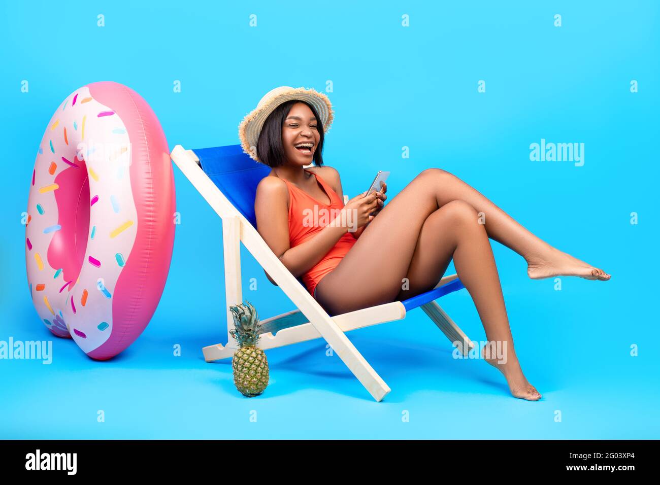Lovely black woman relaxing in lounge chair, using smartphone, chatting to friend online on beach, blue background Stock Photo