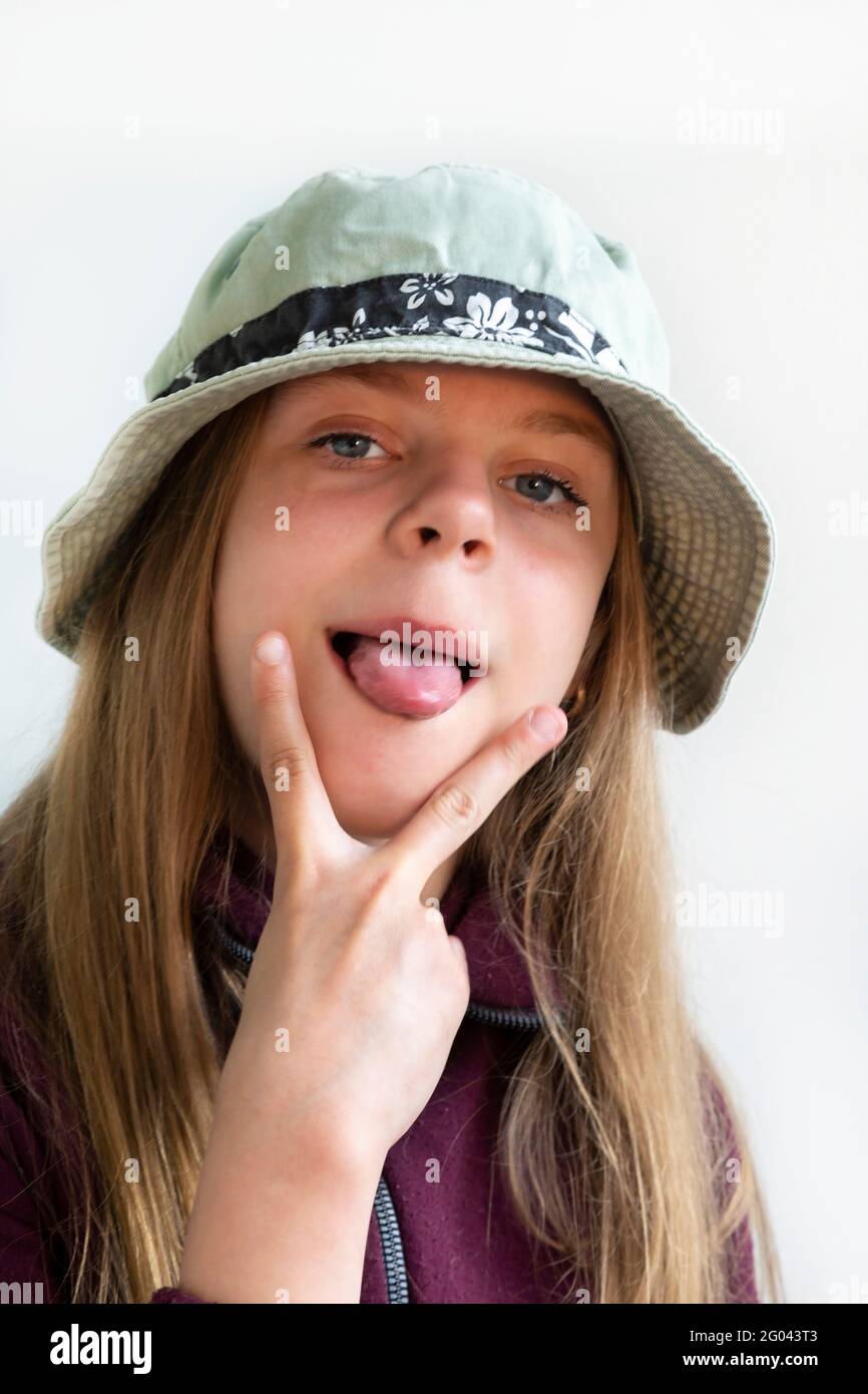 The girl stuck out her tongue and shows two fingers. Cheerful girl with long hair in a green hat. Posing in front of the camera. The joys of a child Stock Photo