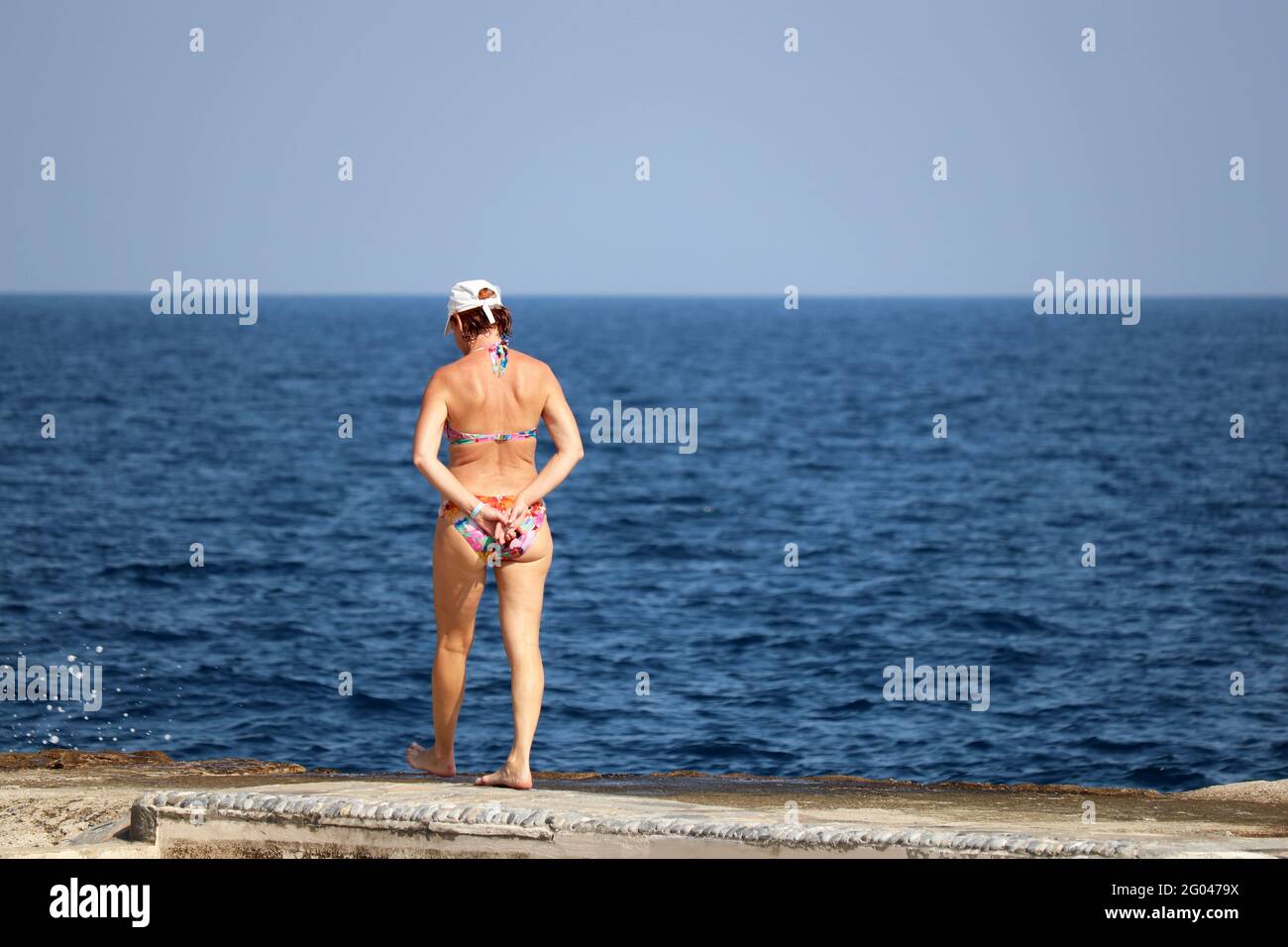 Beach vacation, mature woman in cap and swimsuit walking on stone coast on blue sea background Stock Photo