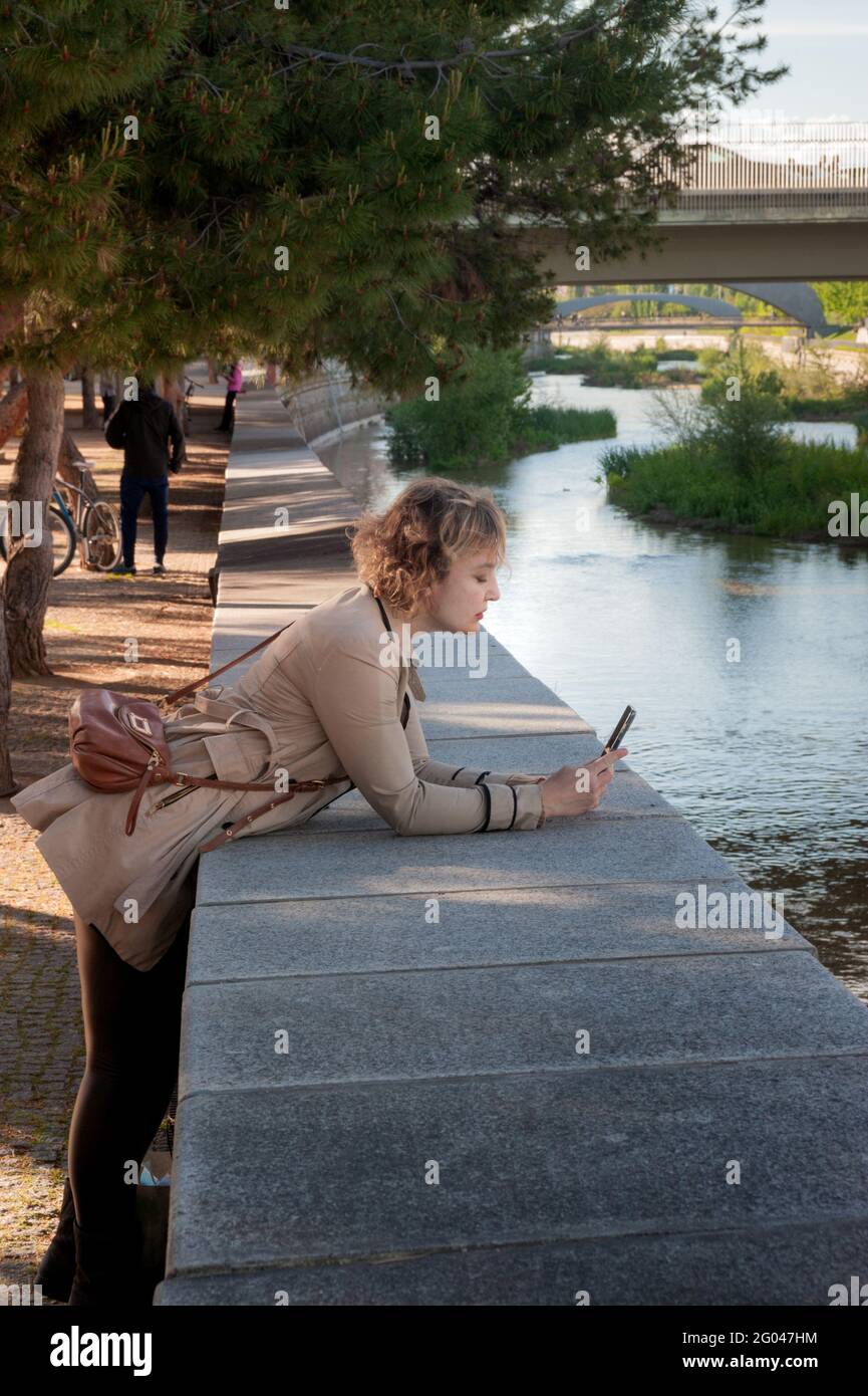 Pretty and elegant middle-aged woman looking her mobile phone in a park leaning against the wall on the bank of a river in the city Stock Photo