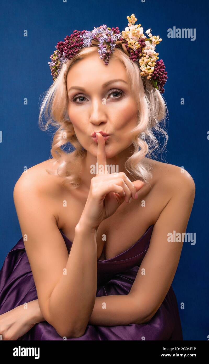 Lovely young beautiful girl in a lilac wreath with a beautiful smile on a blue background, with with daylight, holds finger at mouth Stock Photo