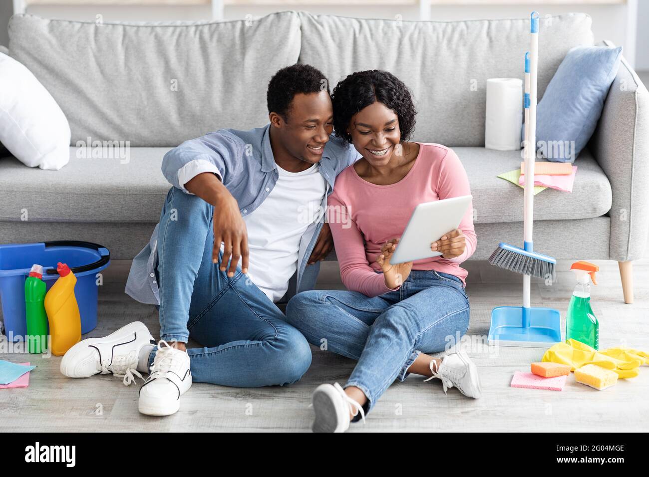 Smiling black couple with cleaning tools resting, using digital tablet Stock Photo