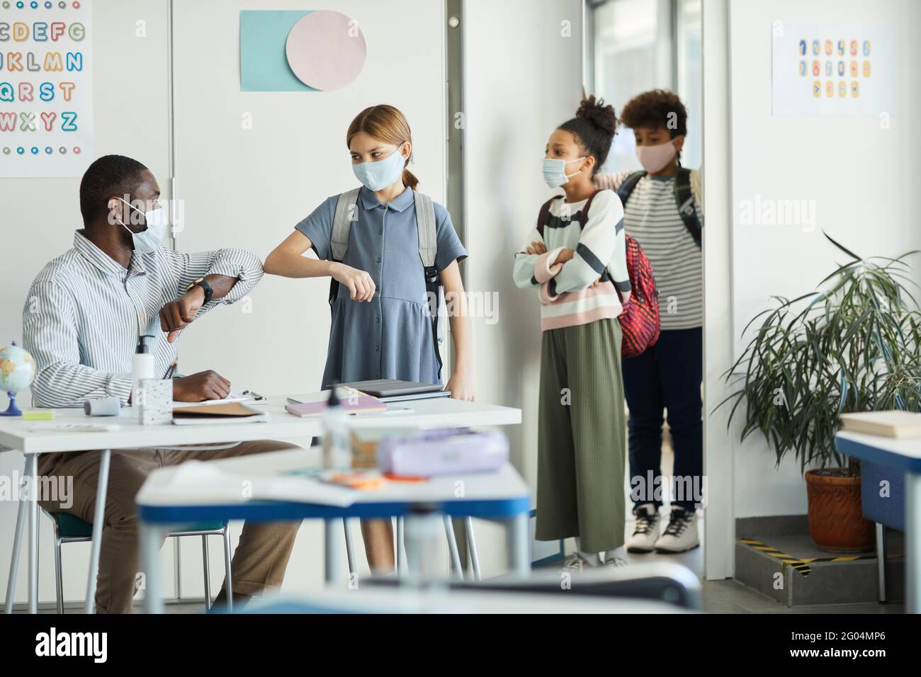 Portrait of male teacher wearing mask and greeting kids entering school classroom, covid safety
