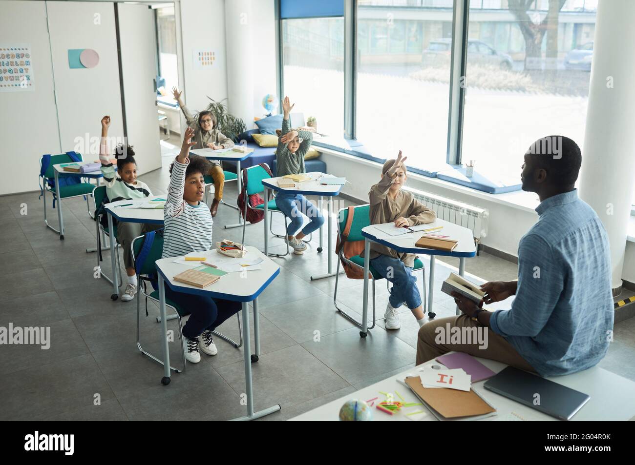 Diverse group of children raising hands in school classroom while sitting at desks, copy space Stock Photo