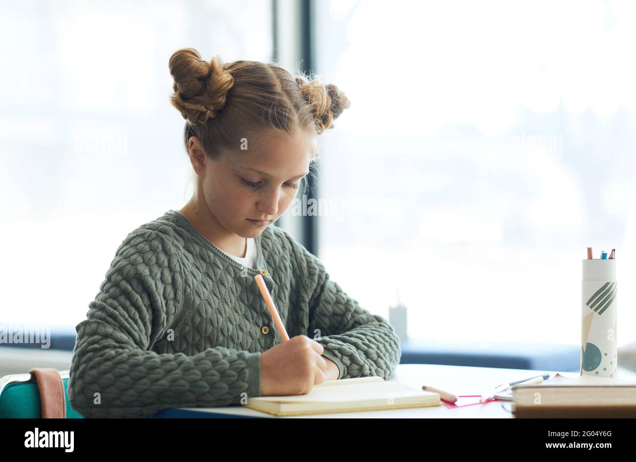 Portrait of cute teenage girl writing in notebook while sitting at desk in school Stock Photo