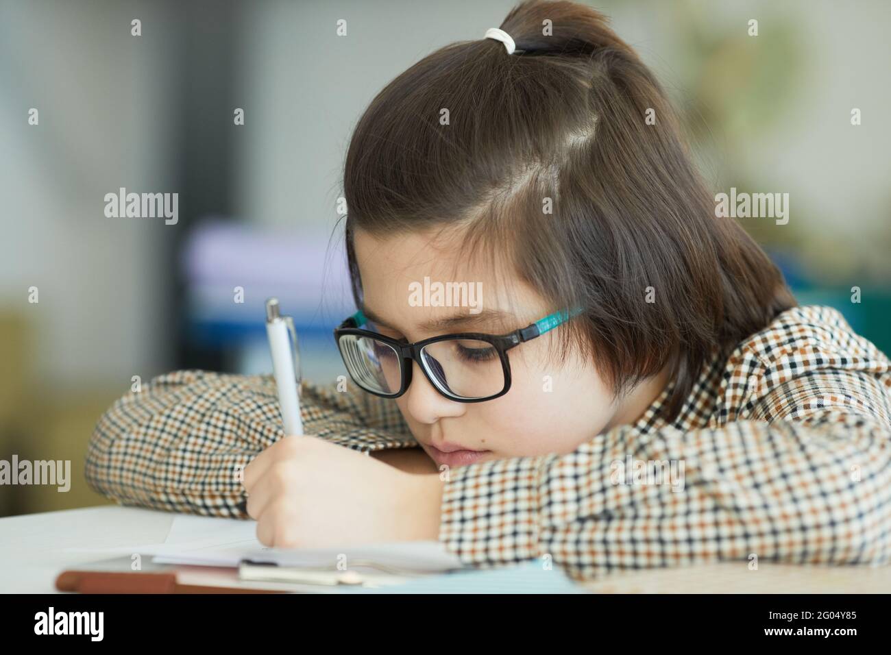 Close up portrait of cute boy wearing glasses sitting at desk in school classroom and writing Stock Photo