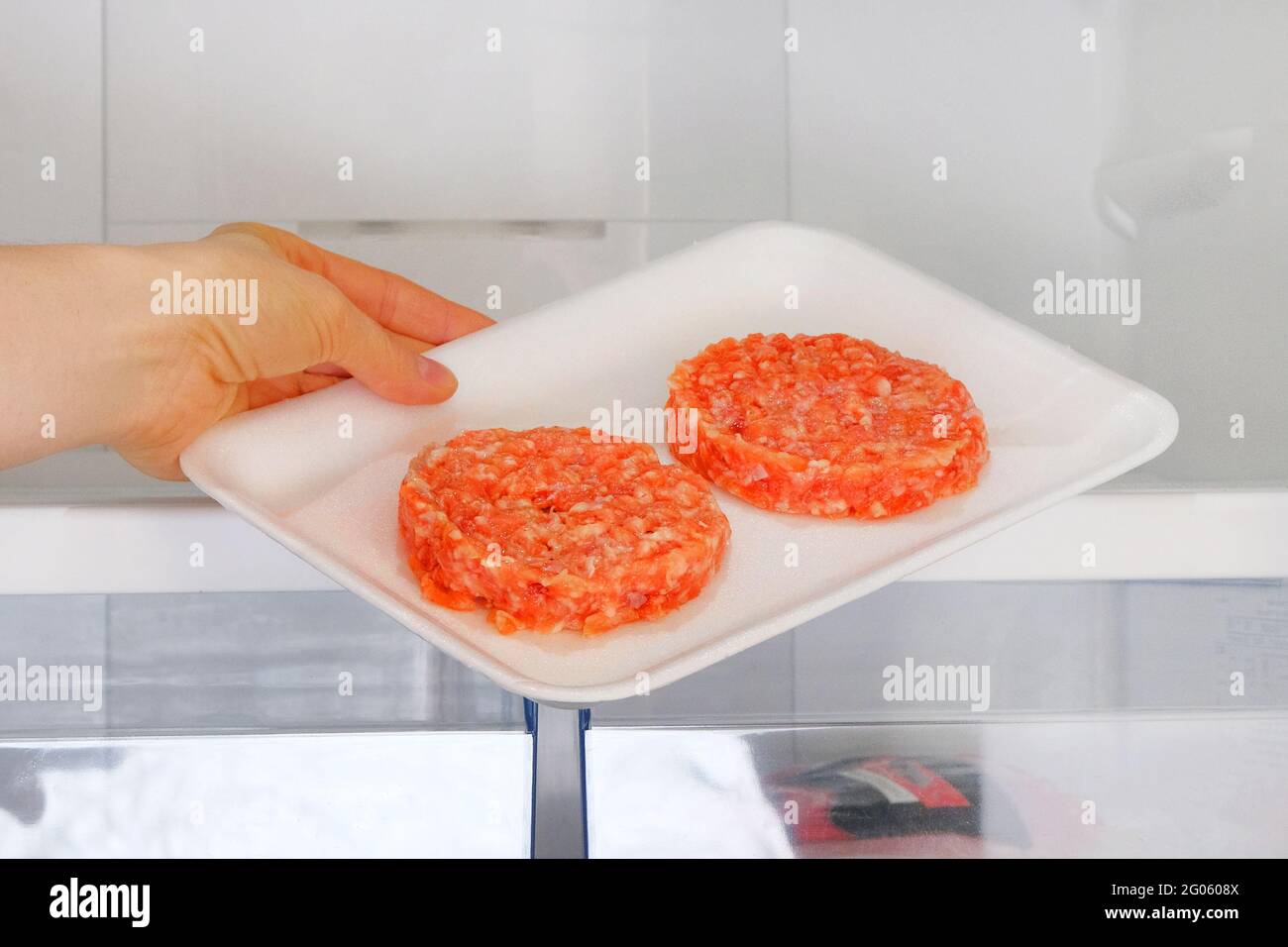 Raw Chicken Burger in container is on shelf in open fridge. Female hand