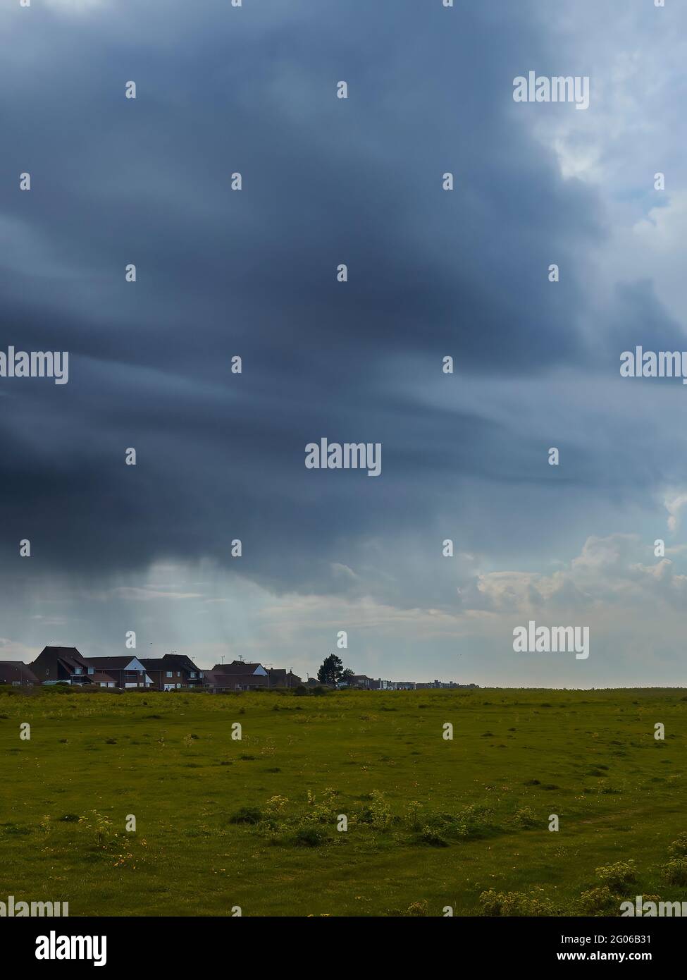 A distant inky storm cloud, hurling rain onto a group of houses, advances to obscure a summery sky with sunlit fluffy clouds, over an empty field. Stock Photo