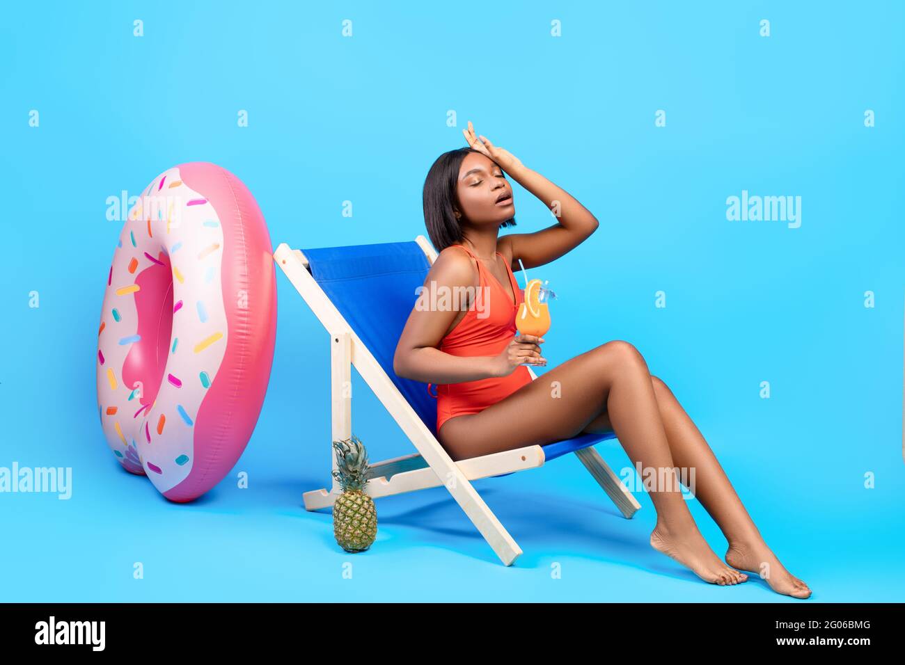 Full length of young black lady suffering from summer heat, holding tropical cocktail while sitting in lounge chair Stock Photo