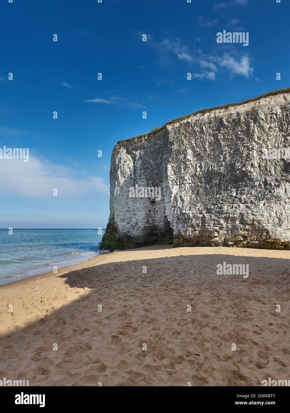 Gentle waves roll in from a blue sea to break on golden sand under a white chalk cliff with a summery blue sky with fluffy white clouds overhead. Stock Photo