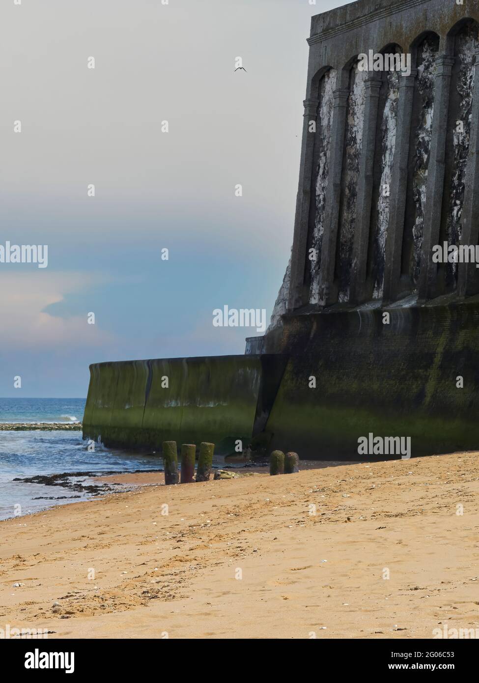A view along a golden sandy beach to some colossal, cyclopean pillars embedded in a chalk cliff with a blue sky bearing a soaring bird behind. Stock Photo