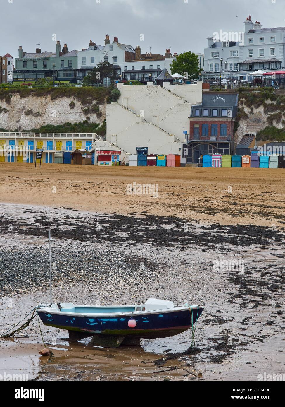 The beach huts and buildings of a cliff-top seaside town. A small boat lies on the sand and a leaden sky hangs overhead. Stock Photo