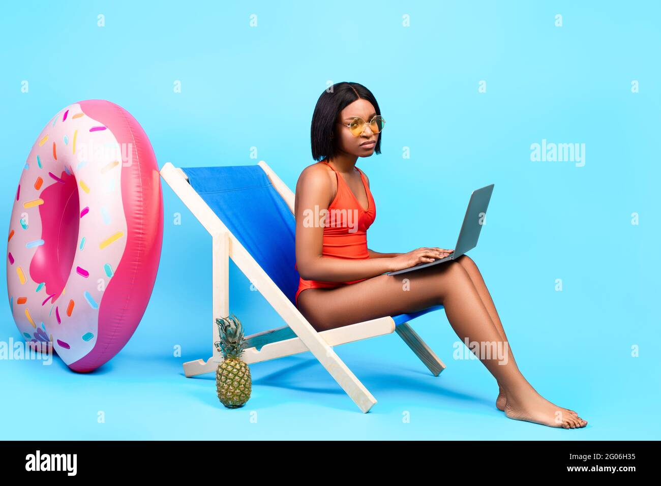 Online work during summer vacation. Unhappy black lady sitting in lounge chair with laptop, having to do job on holidays Stock Photo