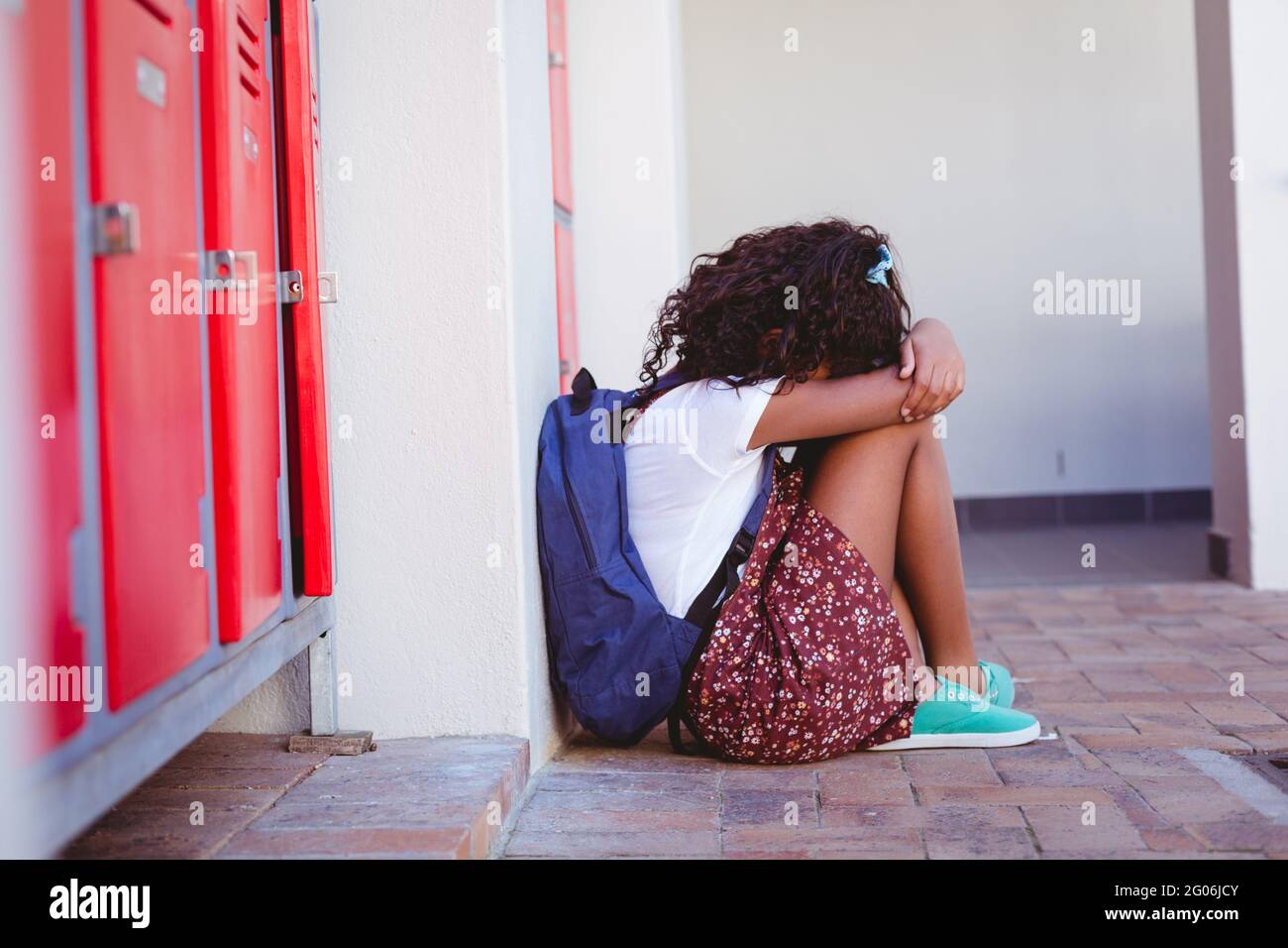 Unhappy african american schoolgirl sitting by lockers in school corridor with schoolbag Stock Photo