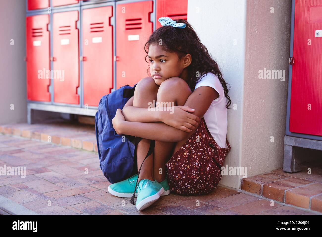 Unhappy african american schoolgirl sitting by lockers in school corridor with schoolbag Stock Photo