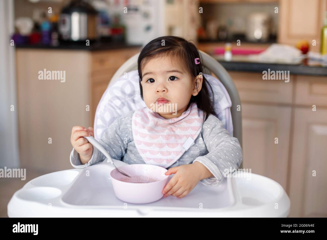 Cute adorable Asian Chinese kid girl sitting in high chair eating soup with spoon. Healthy eating for kids children. Toddler eating independently in k Stock Photo