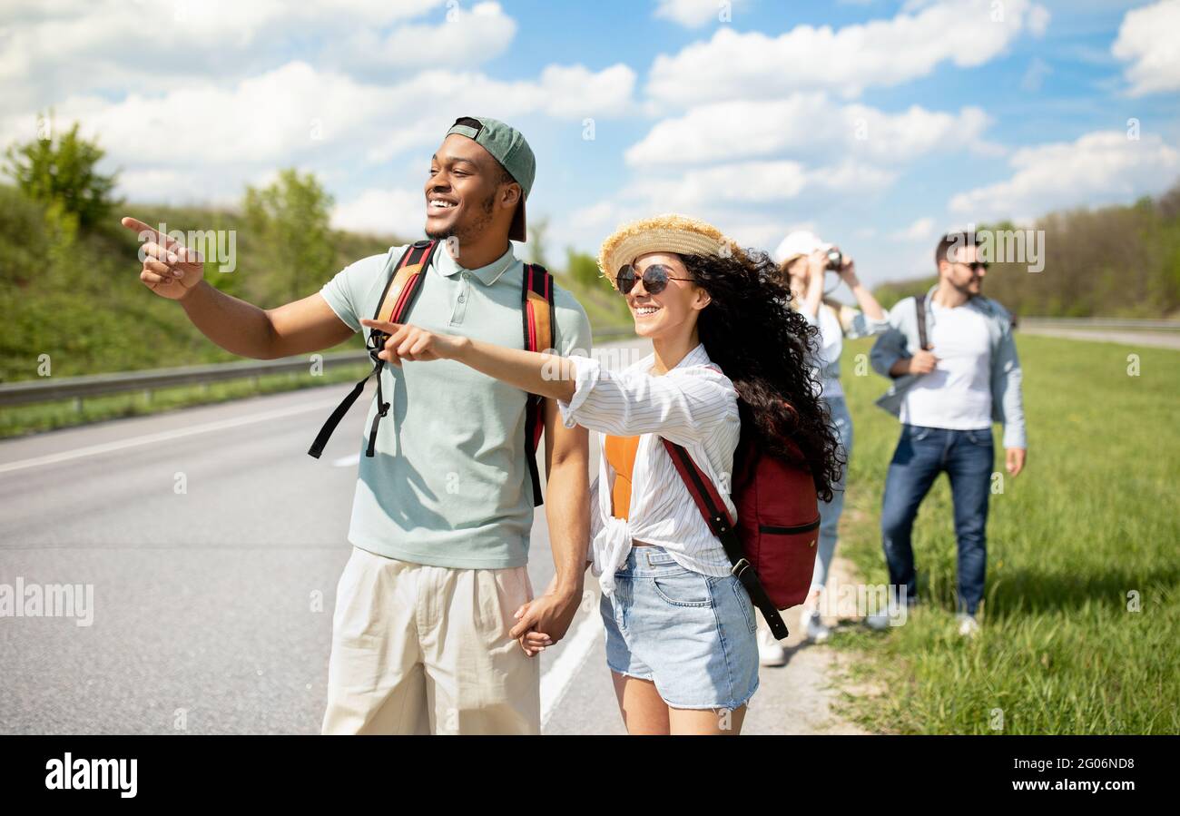 Millennial travelers with backpacks walking along road, hitchhiking in countryside. Group of people having autostop trip Stock Photo