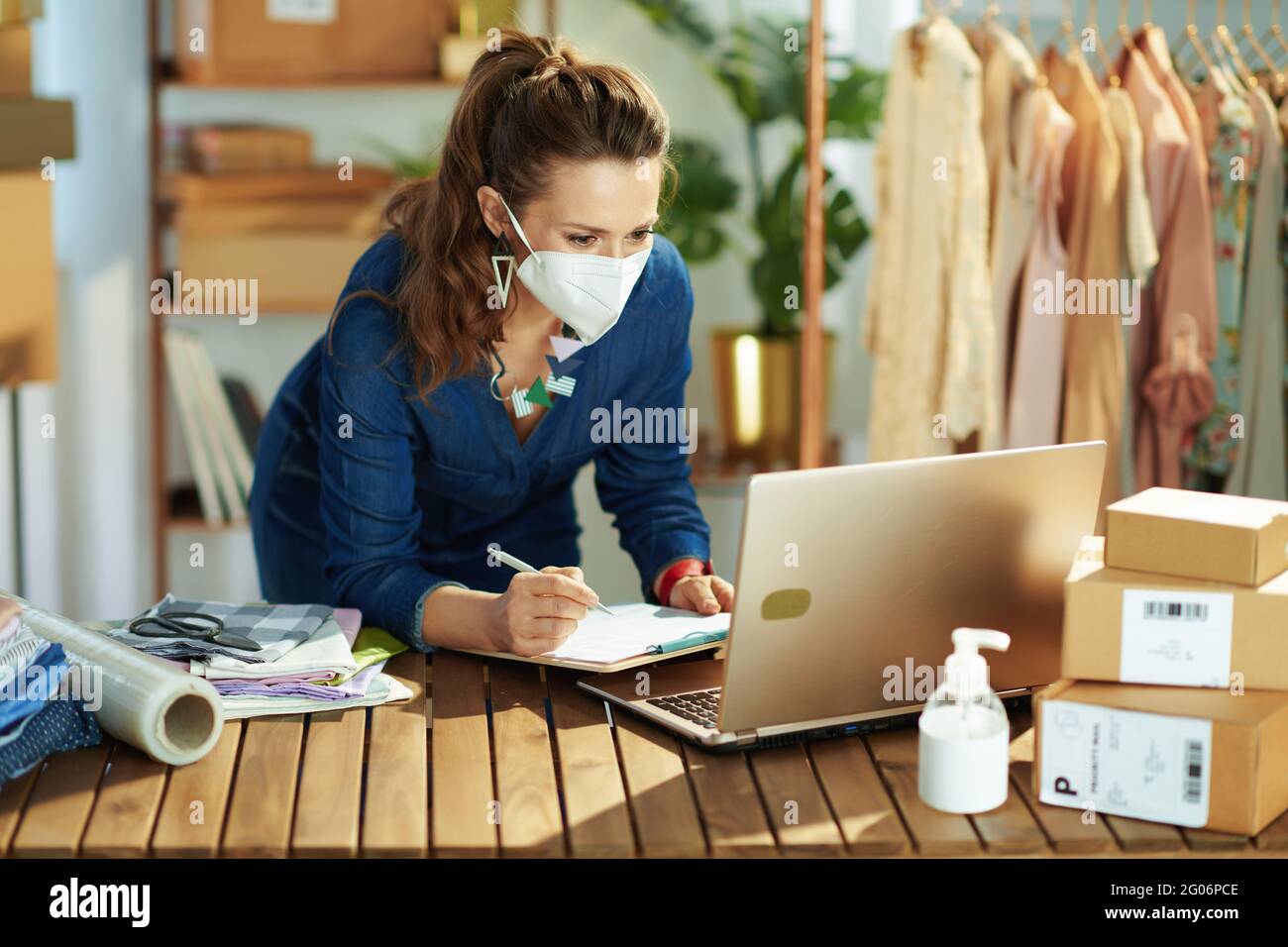 modern small business owner woman with ffp2 mask, parcel and antiseptic using laptop in the office. Stock Photo
