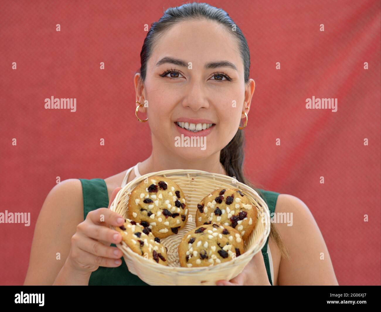Young charming brunette salesgirl smiles to the viewermand shows a basket with home baked gourmet chocolate chip cookies in front of a red backdrop. Stock Photo