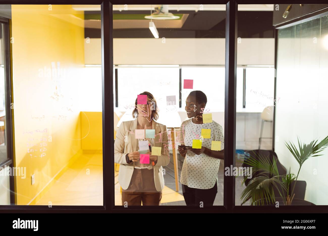 Two diverse businesswomen standing at transparent board, making notes and talking Stock Photo