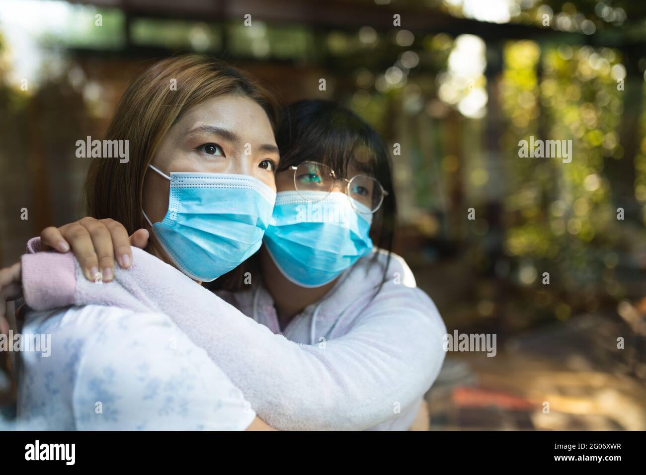 Sad asian woman and her daughter embracing wearing face masks and looking out of window Stock Photo