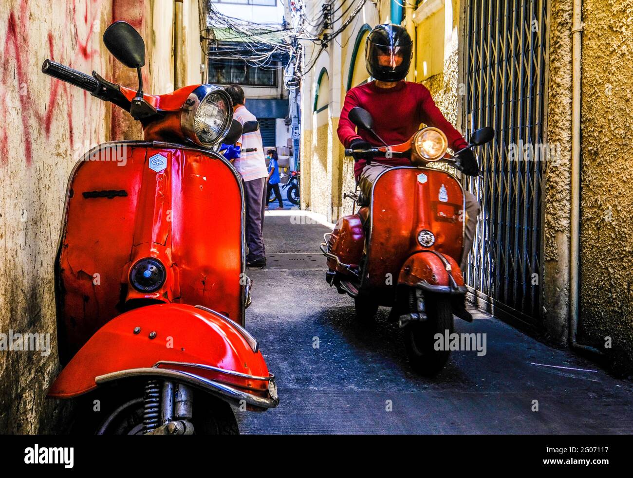 A young male on a red Vespa scooter travels down a narrow alley in Chinatown, Bangkok, Thailand. Stock Photo