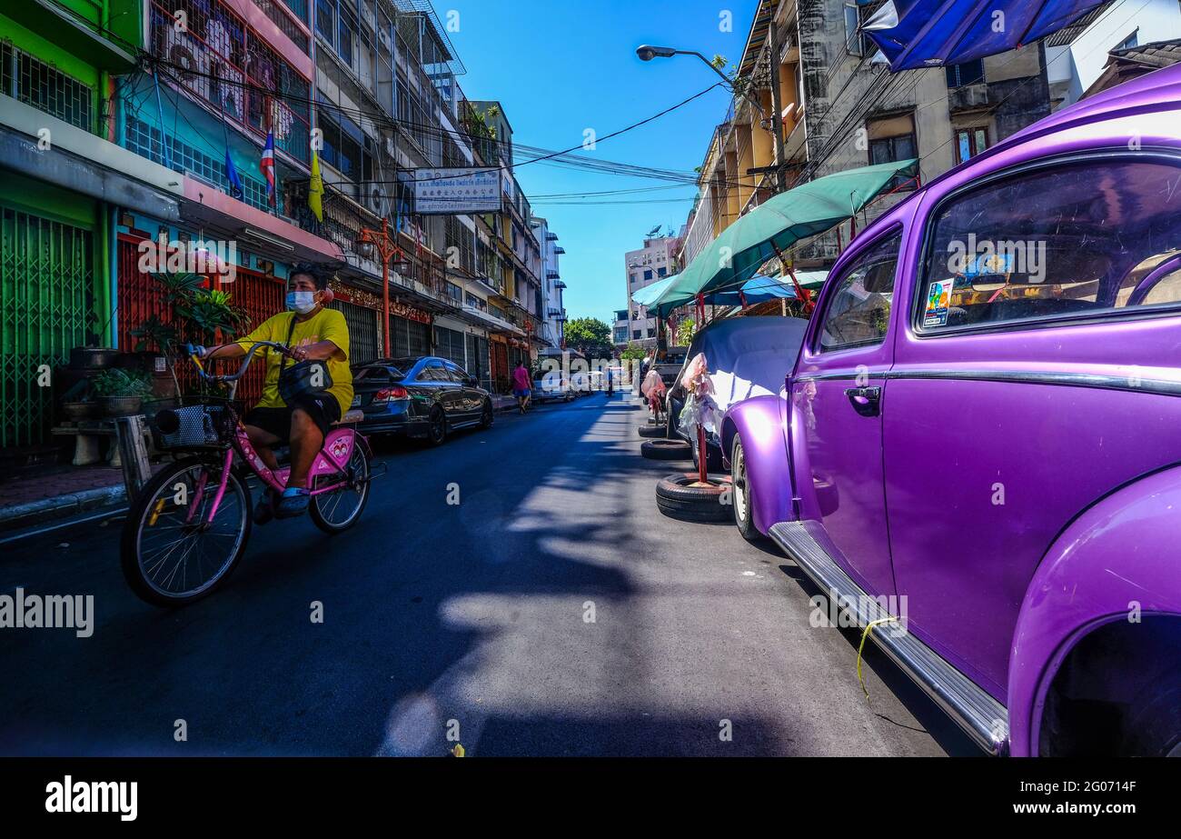 A female cyclist rides past a parked old purple colored VW Beetle against a backdrop of colorful buildings in the Chinatown area of Bangkok, Thailand Stock Photo