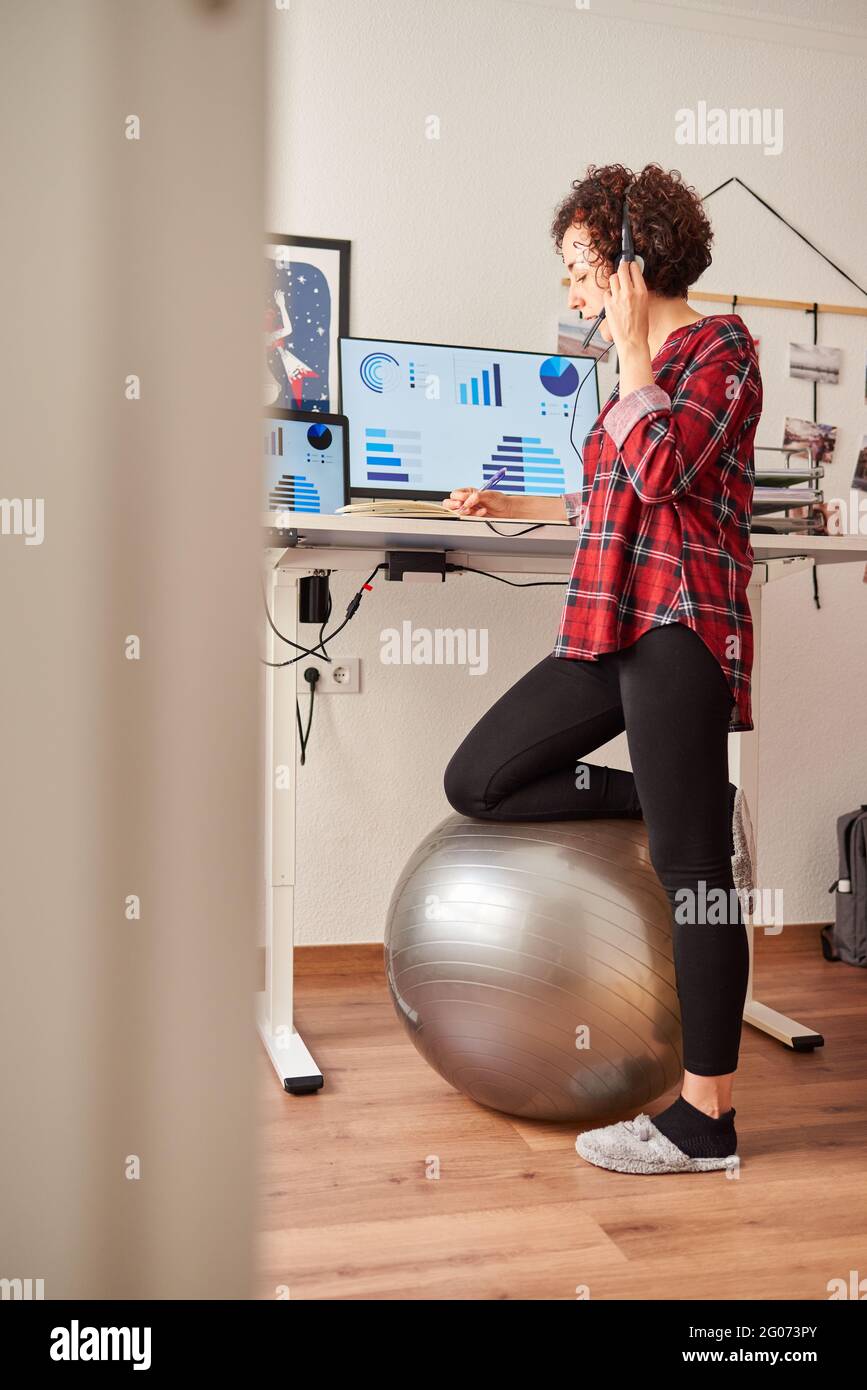 Woman working standing from home with an adjustable height desk and resting her knee on a fitball Stock Photo