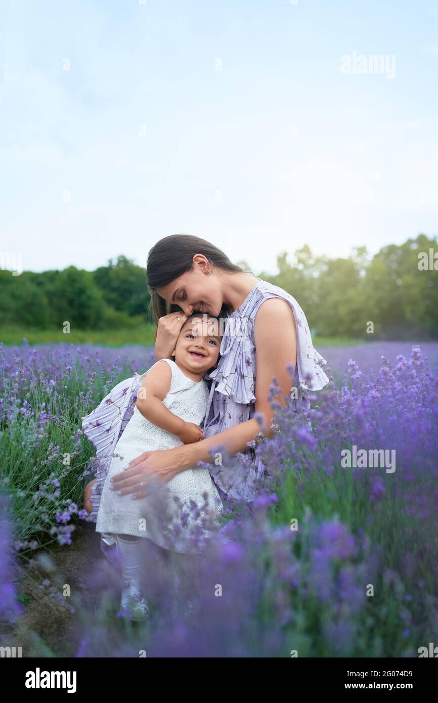 Side view of young mother wearing dress sitting between beautiful purple flowers. Woman posing with little smiling baby daughter on knees in lavender field. Concept of nature, motherhood. Stock Photo