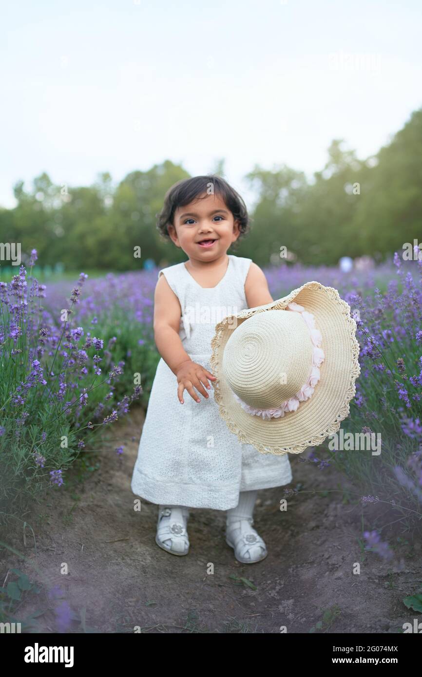 Front view of adorable young female kid posing with straw hat in blooming aromatic lavender field and laughing. Little pretty baby girl wearing lovely white dress enjoying time outdoors. Stock Photo