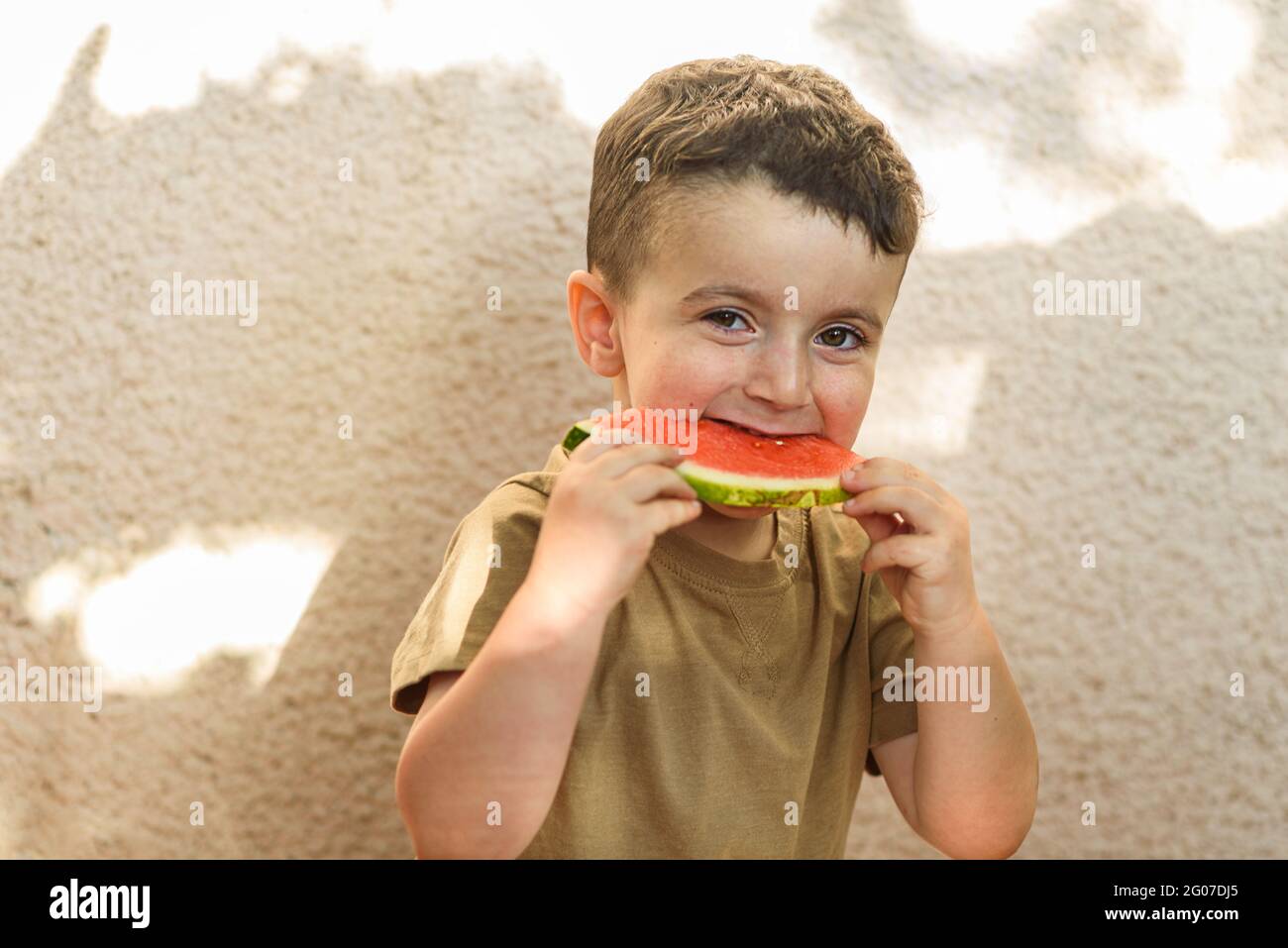 Happy little child eating a slice of watermelon. Boy sits against a white sunny wall in the backyard and bites into a slice of watermelon. Stock Photo