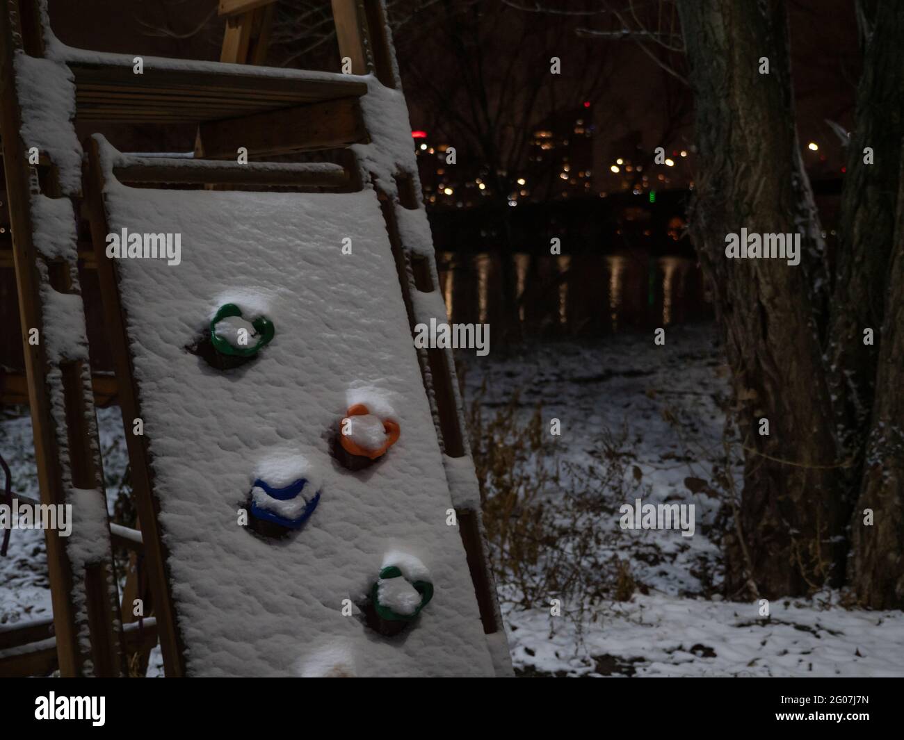 A climbing wall covered by snow at children's playground. Night cityscape with lights reflecting in the river. Winter background. Stock Photo