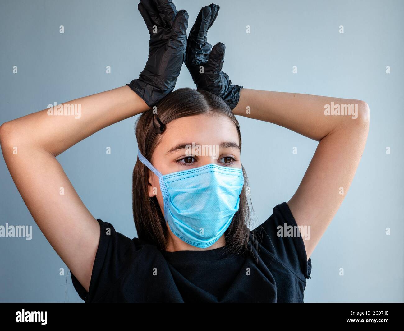 Female teenage girl in a blue protective medical face mask shows corona by holding her hands in black latex gloves above her head. Coronavirus concept Stock Photo