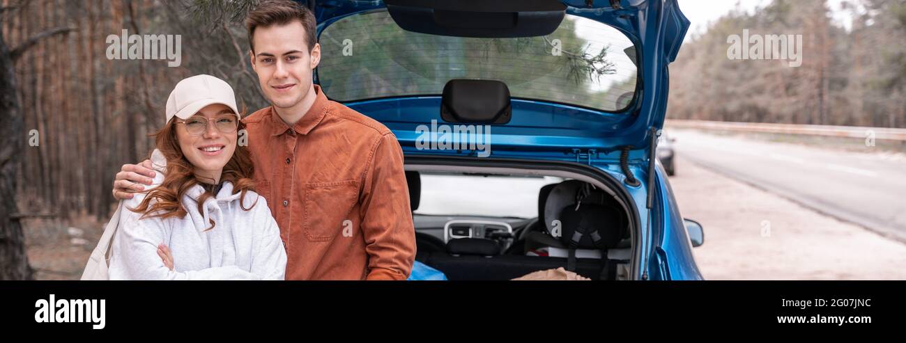 happy man hugging woman in glasses and cap near car in woods, banner ...