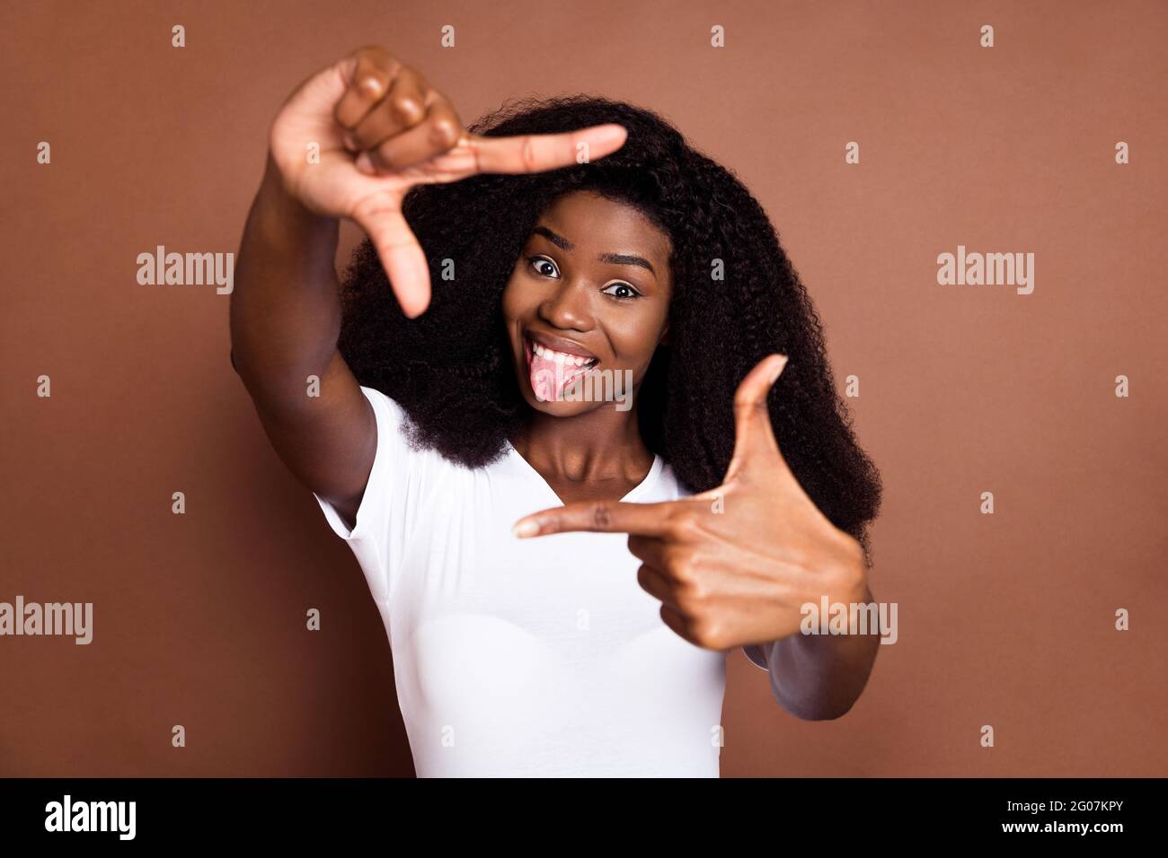 Photo of young black girl happy positive smile grimace tongue-out show fingers frame sign isolated over brown color background Stock Photo