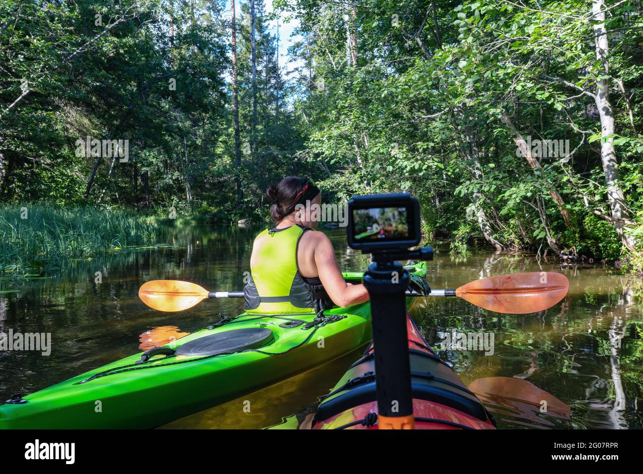Middle age Caucasian Scandinavian women kayaking in small river in forest, one kayak with action camera collides another. Green and red kayaks, sunny Stock Photo