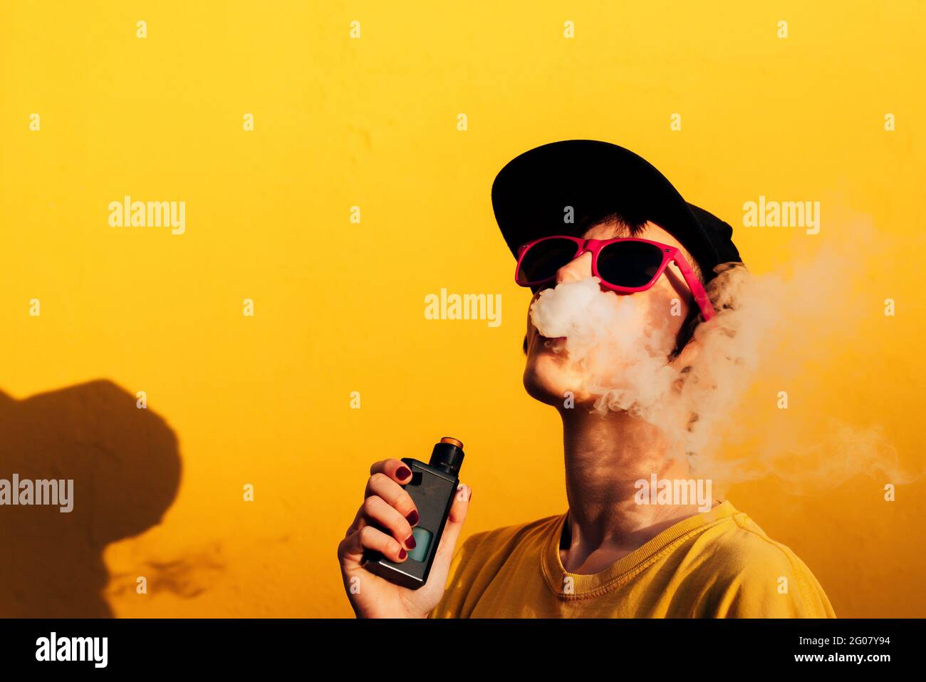 Contemporary female in stylish outfit exhaling fume while standing near yellow wall and vaping on city street Stock Photo