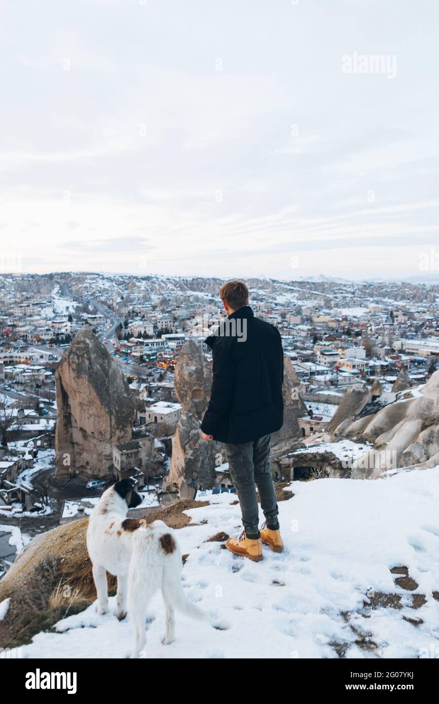 Back view of a young unrecognizable man in warm clothes standing with loyal dog on hill against small ancient cave houses in snowy valley at dusk in T Stock Photo
